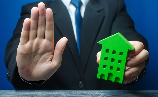 Man with a green house makes a stop gesture. Bank refusal to provide a mortgage loan. Housing problems, unscrupulous dishonest company builder. Confiscation pledged property. Refusal residence permit. man holding right hand out in stop gesture and holding green house in left hand.