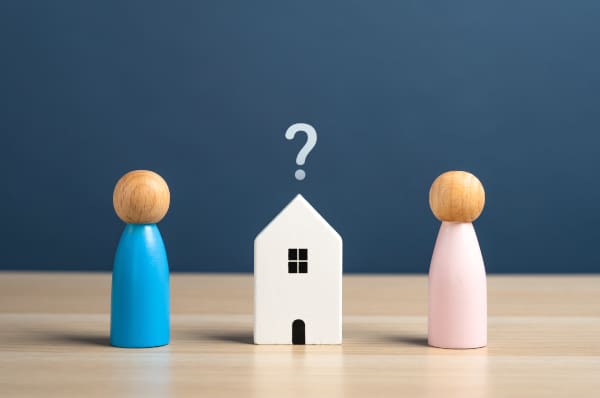 Image of two wooded people standing on either side of a wooden house with a question mark over it indicating why unmarried cohabiting couples must make Wills