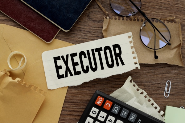 Photo of a desk with numerous office items lying on it and with at piece of town out notebook with the word "excecutor" written on it.