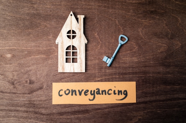 Wooden cutout house sitting on a wooden table with a key and the word "conveyancing" written on a scrap of paper lying on the table
