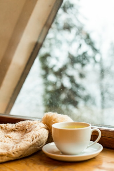 Coffee cup and wooly bobble hat sit on a window sill whilst it snows outside